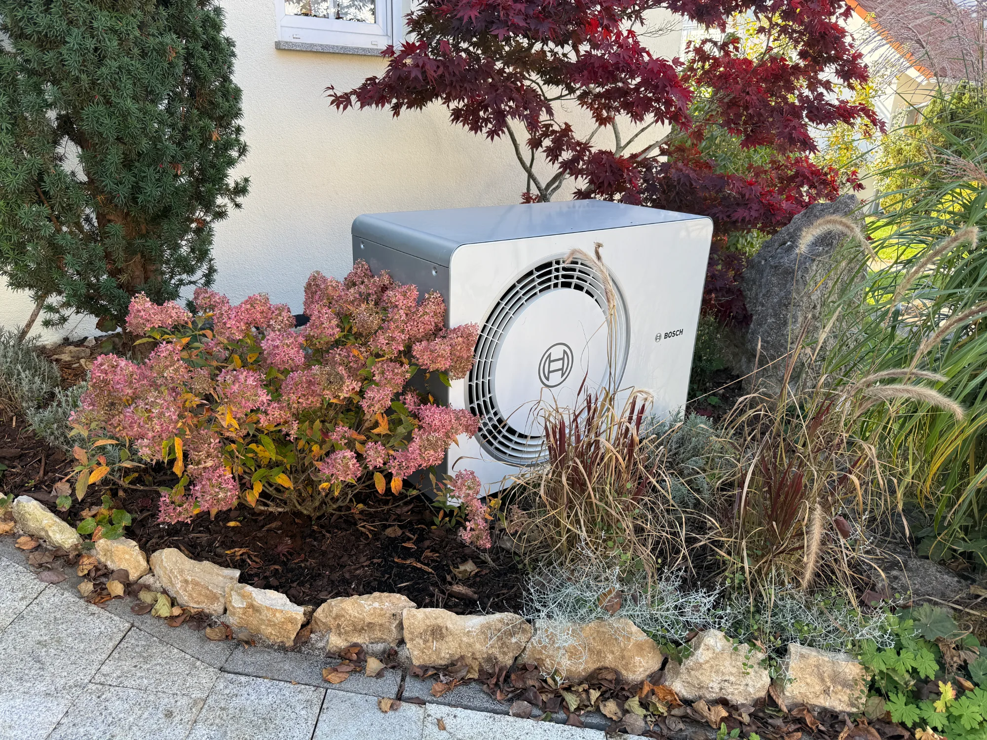 Außeneinheit einer Bosch-Wärmepumpe in einem Gartenbeet vor der Hauswand.