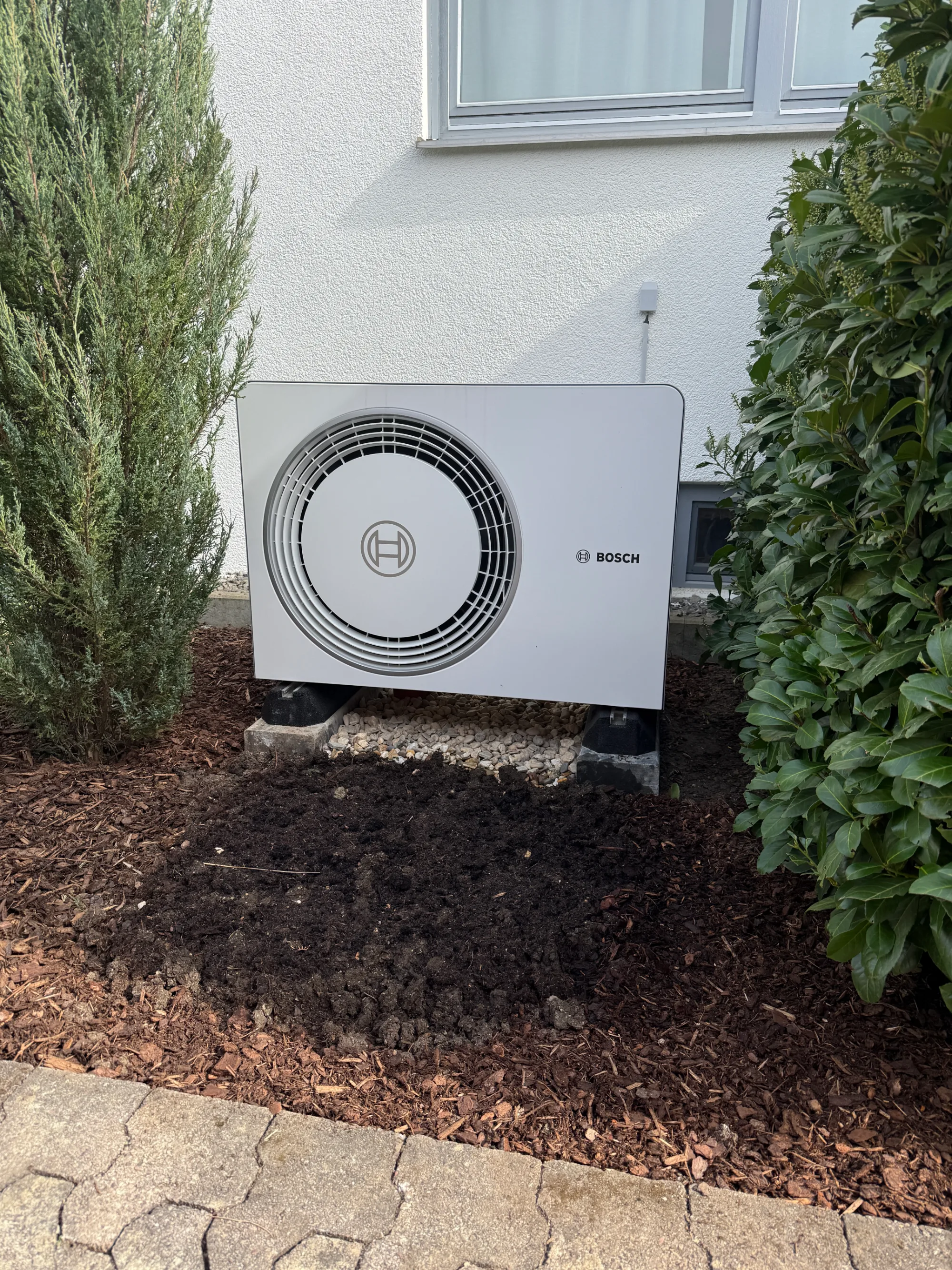 Bosch Außeneinheit einer Wärmepumpe im Beet neben einem Wohnhaus.