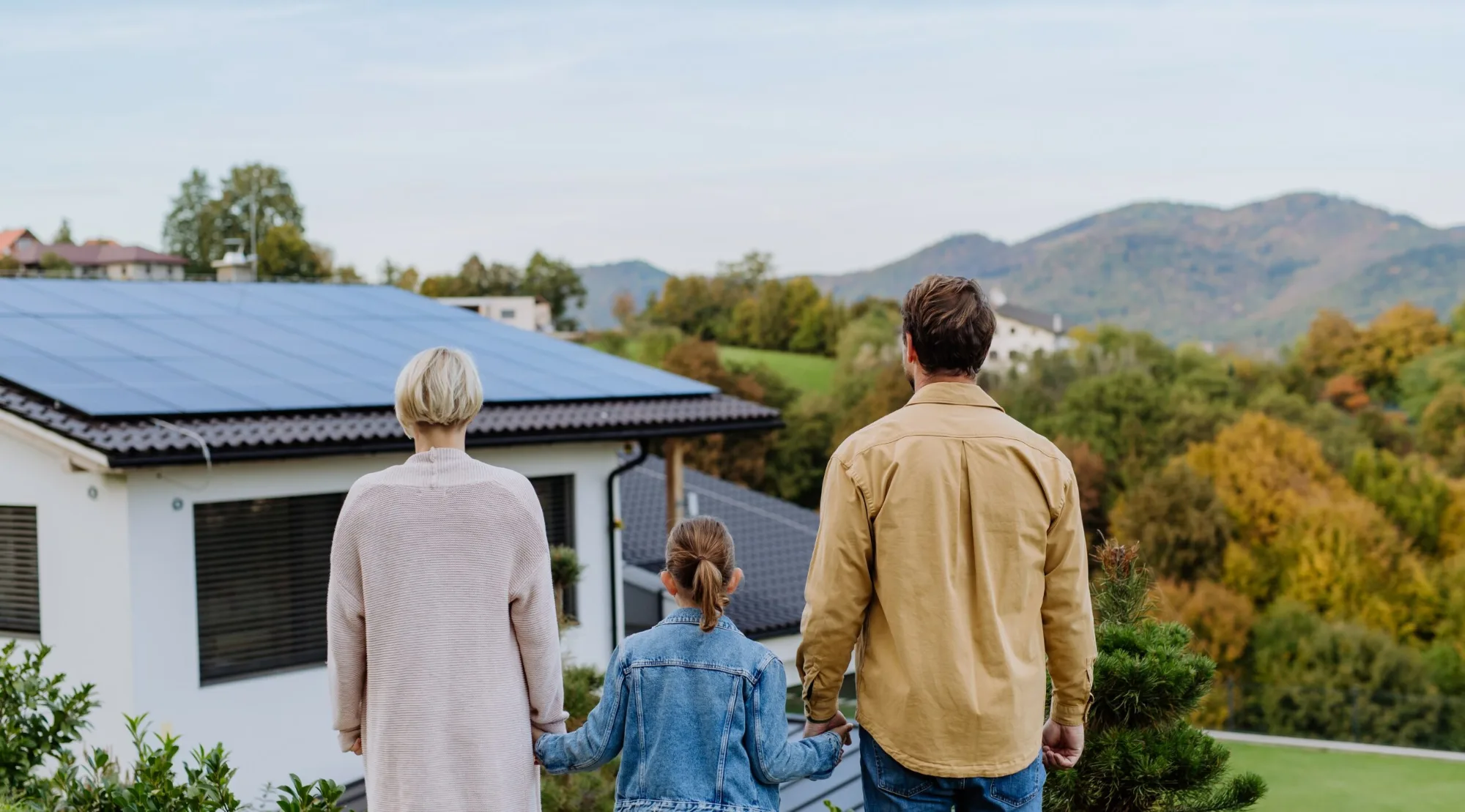 Eine Familie blickt von hinten auf ein Einfamilienhaus mit einer Photovoltaikanlage auf dem Dach.